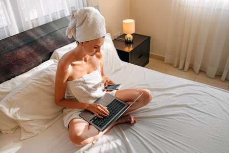 Happy caucasian woman using wireless laptop while resting on bed in bath towel. Female traveler enjoying time at comfy hotel room with modern device.の写真素材