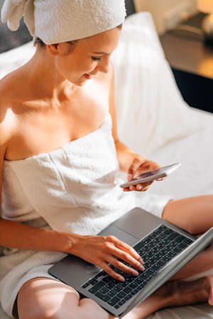 Close up of young woman scrolling with finger on mobile screen while resting on bed with laptop on knees. Female using modern gadgets at cozy hotel room.の写真素材