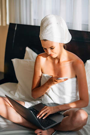 Woman wrapped in a bath towel lying in bed with a laptop on her lap and holding a smartphone in her hands. Lifestyle with modern technologies. Young girl with a laptop talking on the phone and smilingの写真素材