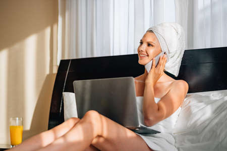 Pretty woman wrapped in bath towel lying on bed with laptop on knees and holding smartphone in hands. Lifestyles with modern technology. A woman works on vacation at the hotel, after a bath or shower.の写真素材