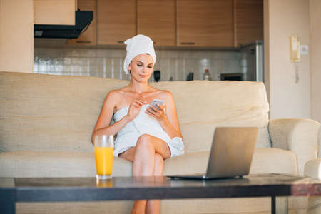 Charming caucasian woman sitting on cozy sofa and opening modern laptop. Pretty female in bath towel surfing internet while resting at bright hotel room.の写真素材