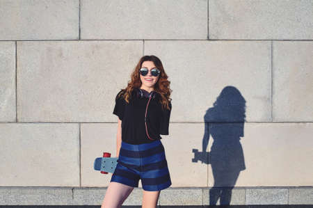 A Girl Stands on the Background of a Gray Wall In The Sunglasses With a Skateboard and Smiles.Hipster Conceptの写真素材