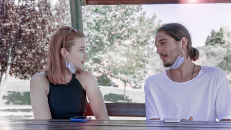 Happy young friends wearing face masks having a conversation while sitting on a wooden bench in the...