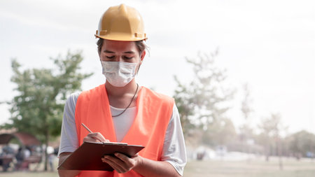 Young male construction worker with yellow helmet and face mask taking notes at construction site. New normality after covid-19 outbreak.の写真素材