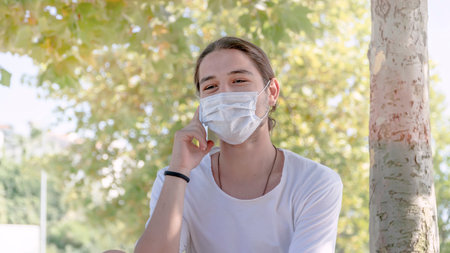 Young happy man with face mask talking on the phone outside. New normal after coronavirus outbreak....