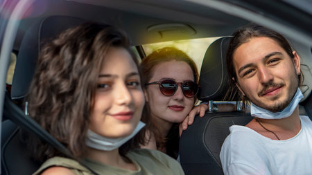 Young friends wearing protective face mask looking at camera are sitting in the car. Corona virus...