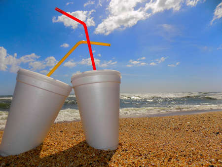 Two glasses of expanded polystyrene (polystyrene foam), with lids and straws, on the beach, partially buried in the sand.の写真素材