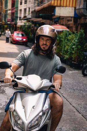 Vertical portrait of mid adult man driving a scooter with an open face helmet in the southeast of Asia; front view in a street of Sukhumvit, Bangkok, Thailandの写真素材