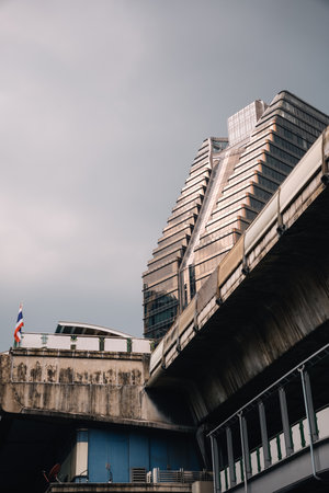 Glassy skyscraper emerging next to a large concrete bridge structure with the reflection of the sun at sunset; Bangkok, Thailandの写真素材