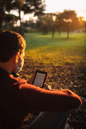 boy with a mask by the covid-19, reading an e-book, sitting in a tree at sunset.の写真素材