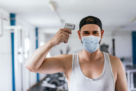 Man with mask taking his temperature. Infrared thermometer. Healthy gym. Pandemic, coronavirus. Concept of health and safetyの写真素材