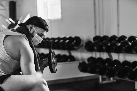 Man with mask doing biceps exercises with dumbbells in the gym. sitting on a bench. Health and wellness conceptの写真素材