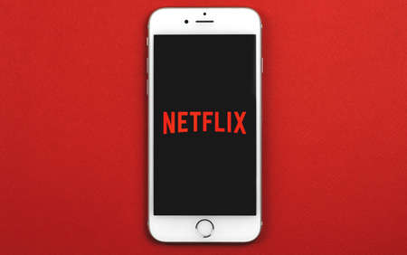 Kharkov, Ukraine - February 22, 2021: Netflix app logotype on screen of Apple smartphone, studio shot, flat lay red background, copy space photoのeditorial素材