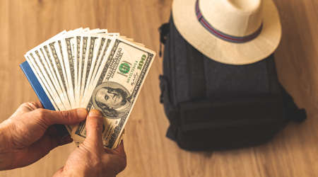 Traveler shows money for future trip, travel preparation concept, man with bunch of US dollars background of bag and straw hatの写真素材