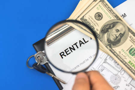 Man search for the rental concept. Rental agreement and magnifying glass in hand. Clipboard with document on the office table with house keys. Blue backgroundの写真素材