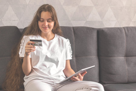 Smiling young caucasian woman with credit card shopping from home sitting on sofa in living room...