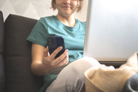 Smiling girl using smartphone and laptop indoors in autumn weekend day, hand with modern mobile...