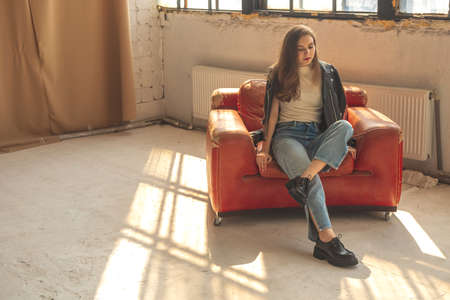 Girl sitting on old leather armchair in studio interior, casual and urban clothes and fashionの写真素材