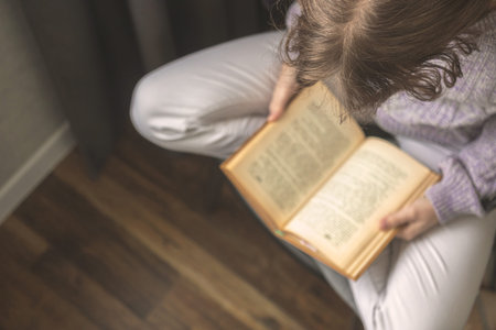 Young woman reading book while sitting in lotus pose on chair in cozy living room, concept of...