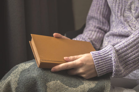Senior woman open book while sitting on chair in living room at cold winter days