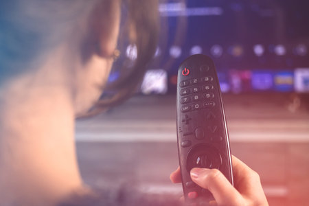 Young woman with TV remote control in hand watching television sitting on sofa at modern living...
