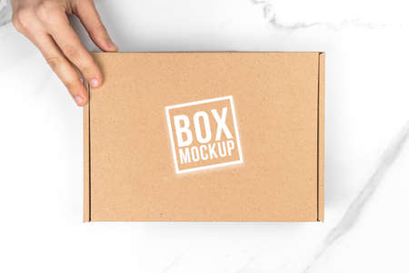Cardboard box mockup in delivery man hand, top view photo. White table background. Suitable for food, cosmetic or medical packaging. Shipping and delivery conceptの写真素材