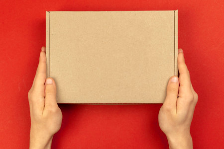 Delivery man hands with cardboard box mockup, top view. Christmas gifts concept. Red table background. Shipping and delivery, copy spaceの写真素材