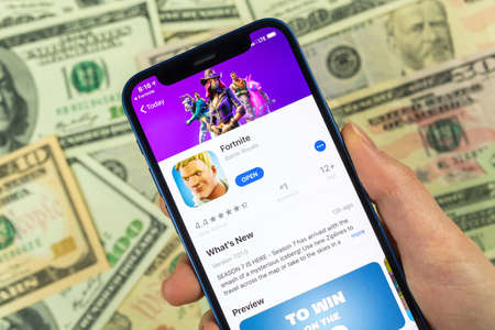 Kharkov, Ukraine - January 14, 2022: Person uses Apple iPhone for playing Fortnite game. Icon close-up, money, dollar bills business backgroundのeditorial素材