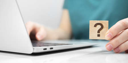 Customer care and support concept background. FAQ question mark icon on a wooden cube close-up view, laptop on a white office table photoの写真素材