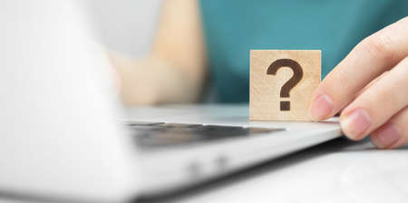 Customer care and support concept background. FAQ question mark icon on a wooden cube close-up view, laptop on a white office table photoの写真素材