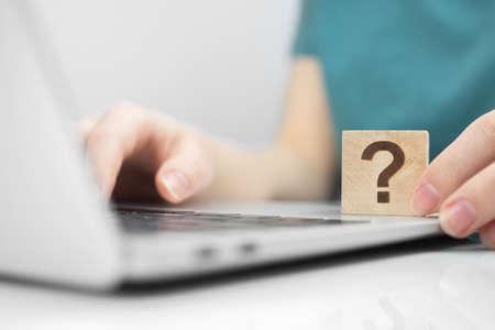 Customer care and support concept background. FAQ question mark icon on a wooden cube close-up view, laptop on a white office table photoの写真素材