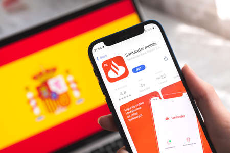 Poltava, Ukraine - April 7, 2022: Santander mobile banking app in Spain. Hand with smartphone, on the screen. background with national flag photoのeditorial素材
