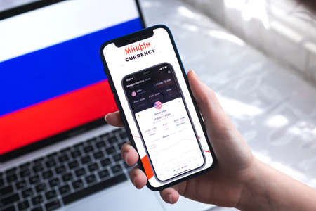Poltava, Ukraine - April 7, 2022: Minfin currency mobile app. Hand with smartphone, on the screen. background with national russian flag photoのeditorial素材