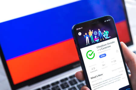 SPB, Russia - April 7, 2022: Sberbank mobile banking app in Russia. Hand with smartphone, icon on the screen. background with national flag photoのeditorial素材