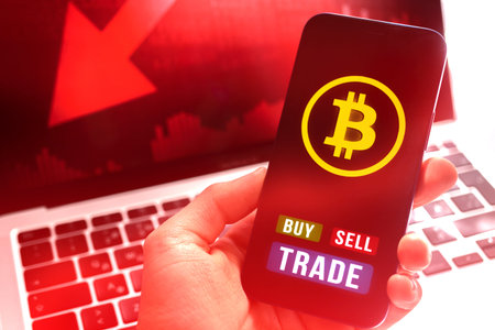 Businessman with bitcoin buy and sell trade application on mobile phone, red stock chart on laptop...