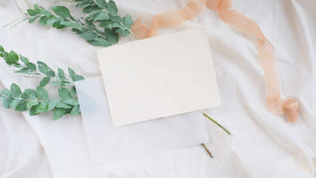 Photostock wedding styled composition. Feminine desktop mockup scene with leaves, silk ribbon, envelope, blank brown greeting card on white textured fabric background. Flat lay, top view.の写真素材
