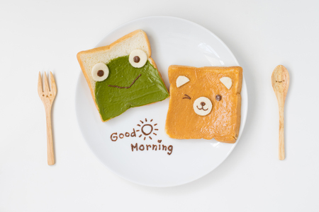 top view of cute breakfast for kids, green tea spread and peanut butter design to frog and bear on breads with good morning text with cute spoon and fork on white backgroundの写真素材