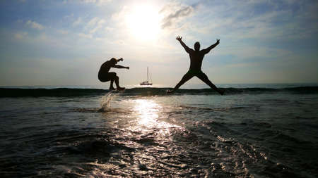 two young men jumping over the waves in the sea, ocean with yacht and sun on the background. Water. Happy. Thailand.の写真素材