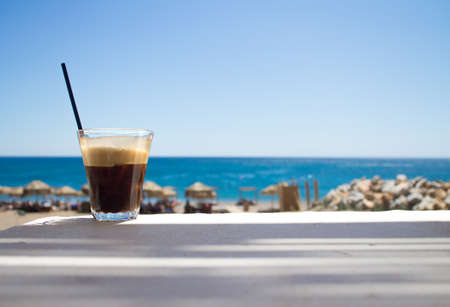 glass with ice coffee fredo espresso or frappe on the corner of the table with beach on backgroundの写真素材
