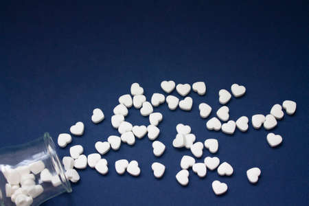 Many of white candy heart-shaped pills spilled out of a transparent jar on a dark blue background.の写真素材