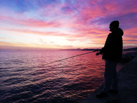 Silhouette of a male fisherman in a hat on the sea embankment with stones, who is fishing against the background of a colorful sunset..の写真素材