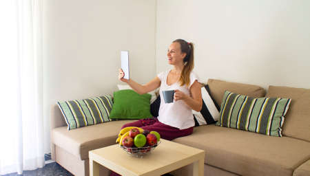 young smiling woman with cup of tea or coffee alone in quarantine self isolated at home and talking on tabletの写真素材