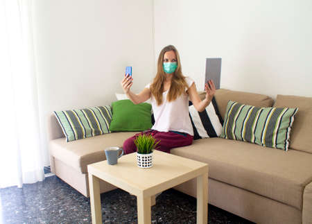 young woman in a medical mask on self-isolation at home is sitting on a sofa with a smartphone and tablet in both handsの写真素材