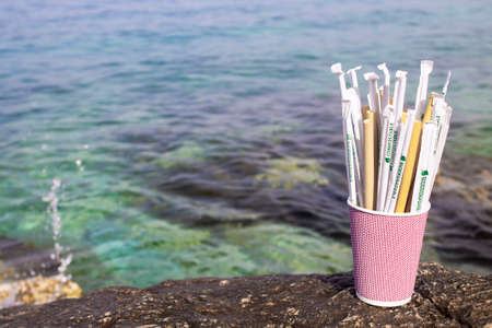 Greece-Athens-20.05.2020. Several replacements of plastic cocktail tubes, bioplastics and bamboo in paper cup on the rocks with blured background with sea. Environment. Saving nature. Bio matireal.の写真素材