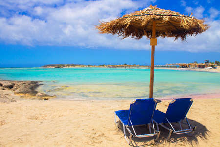 Two blue sun beds and umbrella on pink sand beach with rocks, turquoise water and clouds in blue sky. Wallpaper or background. Tropical view.の写真素材