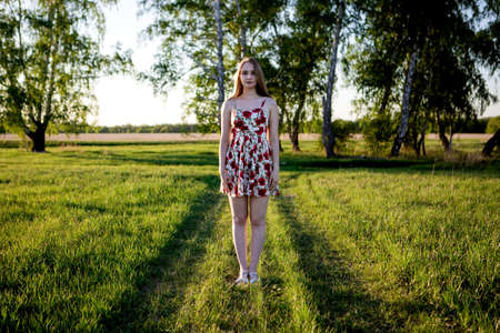 OMSK, RUSSIA, MAY 09, 2020: A girl in a dress on a rural road in a field. A village girl in a short dress with long hair stands on a field road through a field and forest.の写真素材