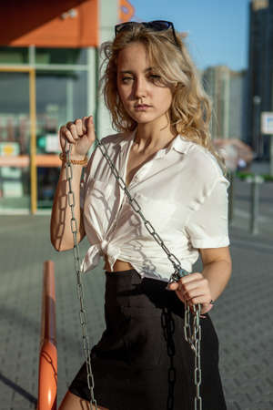A girl in a white shirt poses against the background of an orange building. The blonde in sunglasses holds iron chains with her hands. Portrait of a girl on the streetの写真素材