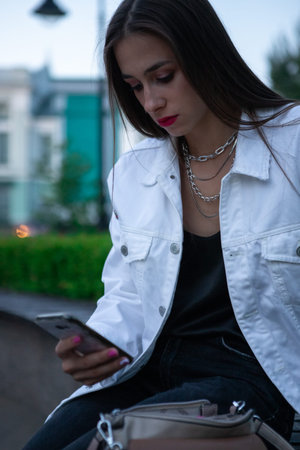 The brunette is sitting on a bench with a phone in her hand. The girl looks at the screen of her...