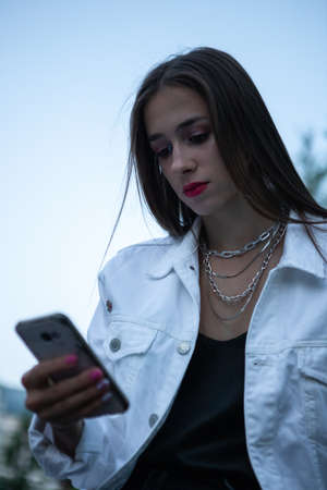 The brunette is sitting on a bench with a phone in her hand. The girl looks at the screen of her mobile phone. Waiting for a message or call.の写真素材