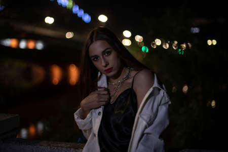 The brunette poses at night against the background of glowing lights along the bridge. Bokeh on the background of a girl. Teen at night in the cityの写真素材